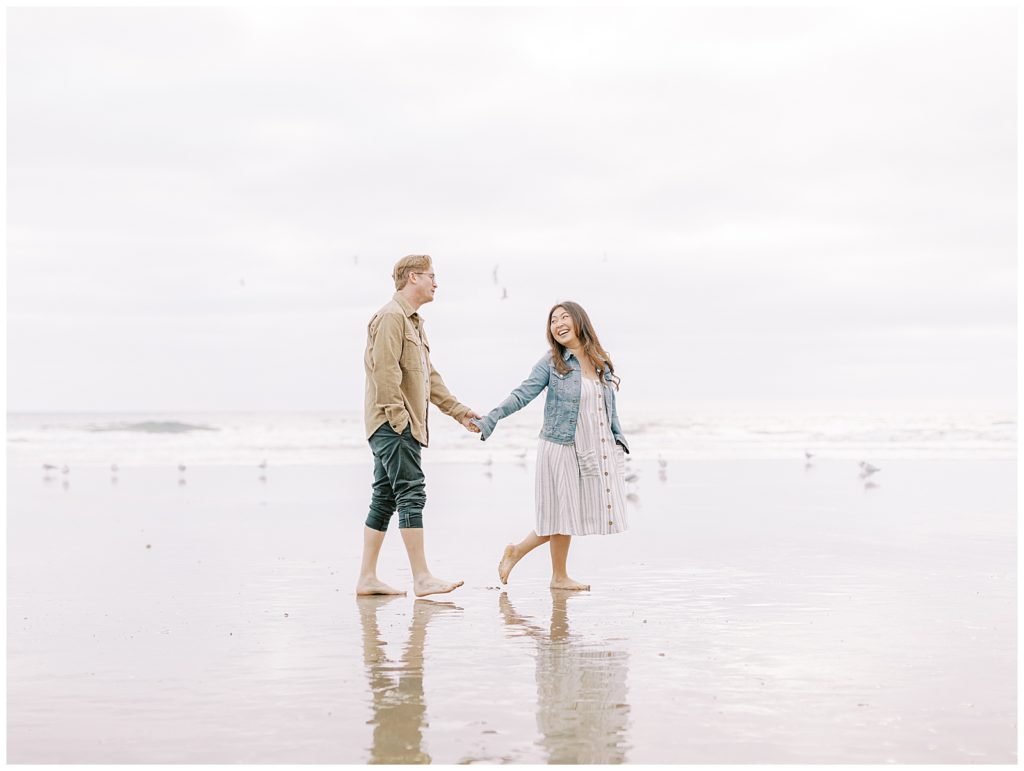 couple on the beach walking towards the ocean holding hands with girl leading the guy