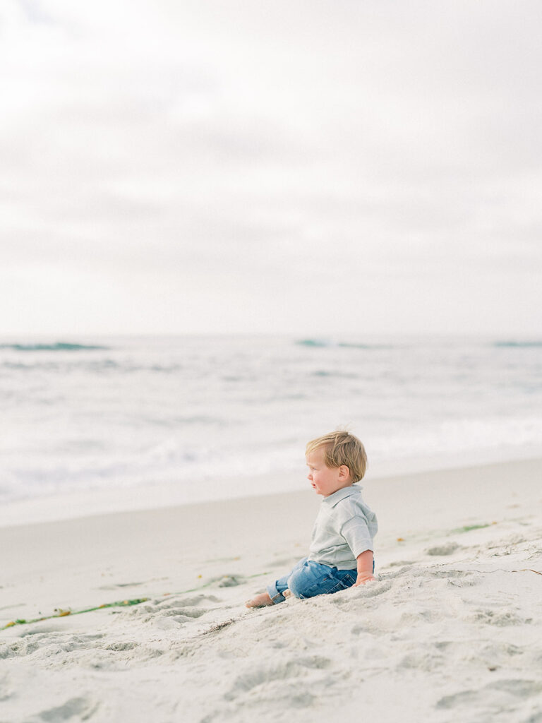 A toddler sitting on the soft sand, gazing intently at the shimmering water as gentle waves roll toward the shore