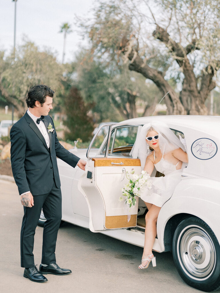 A groom dressed in an elegant suit opens the car door for his bride