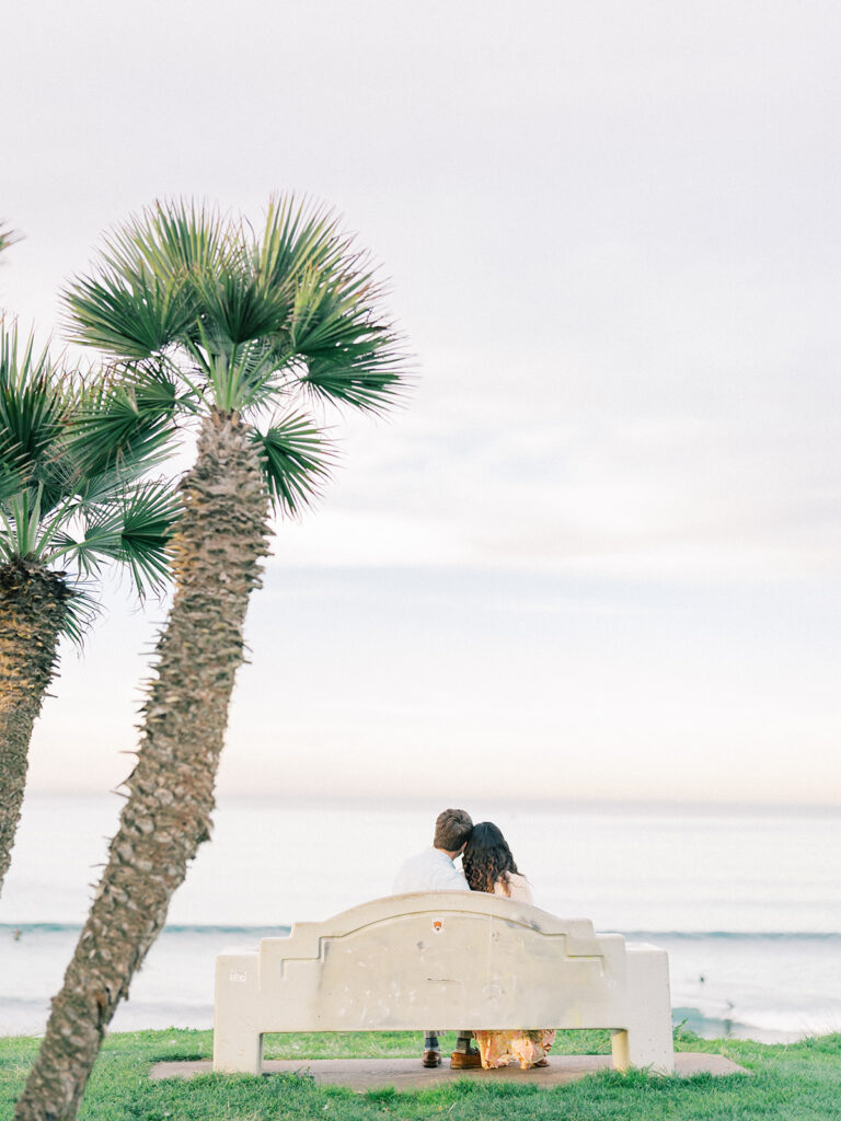 a couple sitting on a bench near palm trees and watching pacific beach captured by san diego beach photographer