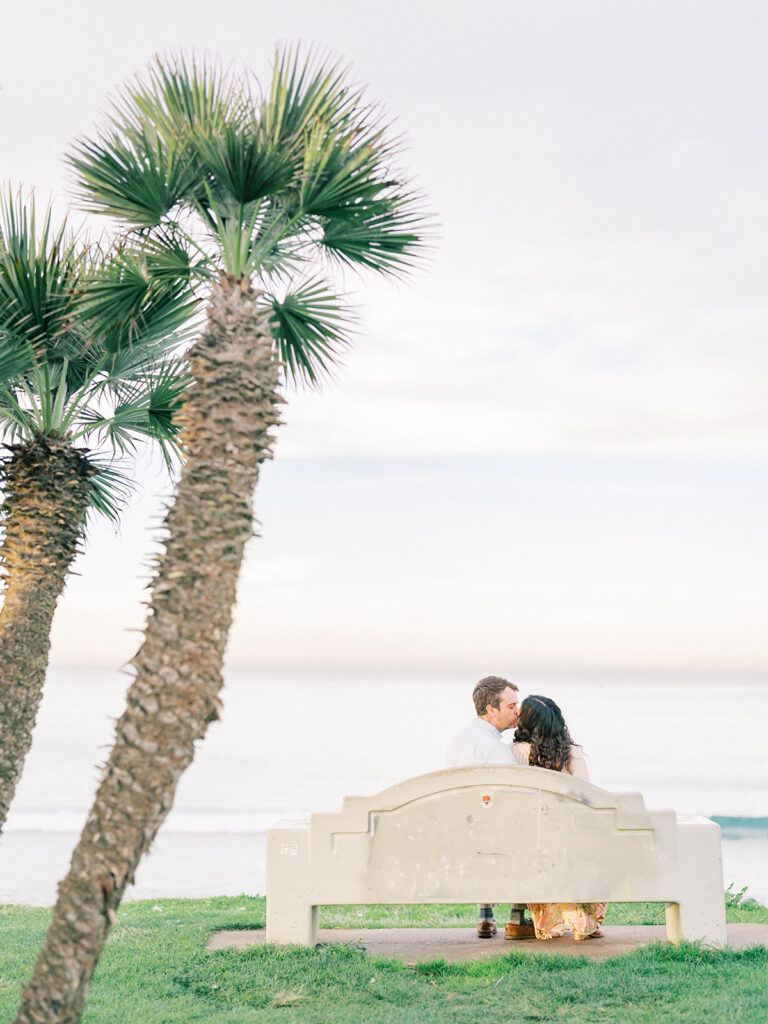 a couple sitting on a bench near palm trees and watching pacific beach captured by san diego beach photographer